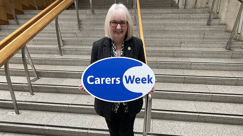 Beatrice stands in the Scottish Parliament holding a sign which reads Carers Week