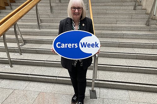 Beatrice stands in the Scottish Parliament holding a sign which reads Carers Week